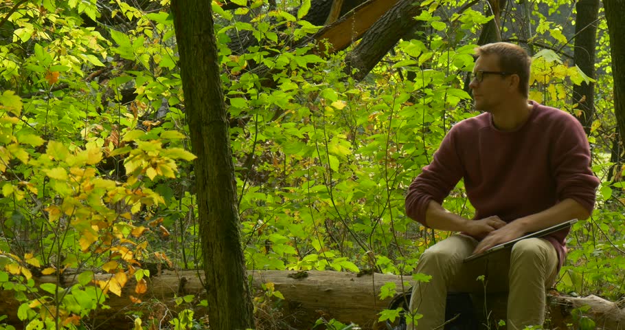 Main in Glasses Sits Comfortably on the Fallen Tree Trunk,Freelancer Programmer Copywriter Designer Accountant Has Pursed His Leg, Opened the laptop, Working with Laptop, old dense forest, fallen