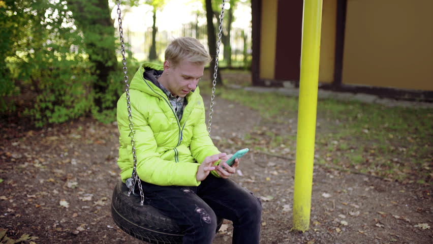 Boy sitting on the swing and browsing internet on smartphone
