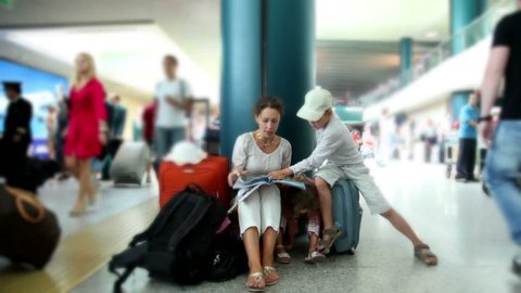 Mother and her two kids boy and girl sit in airport and reading book, their luggage stand near