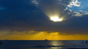 Single masted sailing yacht riding at anchor on a golden horizon. as tourists swim in the tropical sea at sunset. Video UltraHD
