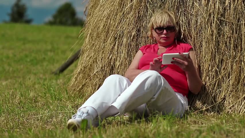 Pretty blond woman sits on green grass near haystack and makes photos on his smartphone