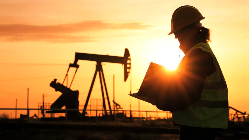 Silhouette of female engineer with clipboard using a cell phone overseeing the site of crude oil production at sunset