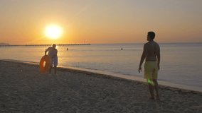 Slow motion clip of man and woman playing with big rubber ring on the beach. They throwing it to each other on the background of sunset over the sea. Summer vacation
