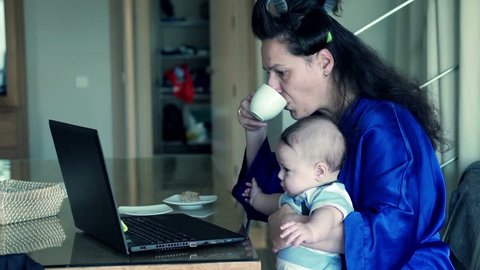 Young, tired mother in bathrobe trying work on laptop with her little child
