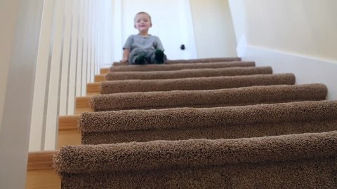 A slow motion shot of an adorable little boy bounces down the stairs while sliding on his bum and laughing
