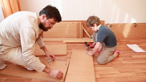 Father tightening screws in wooden board and little boy works with hammer sitting on the floor