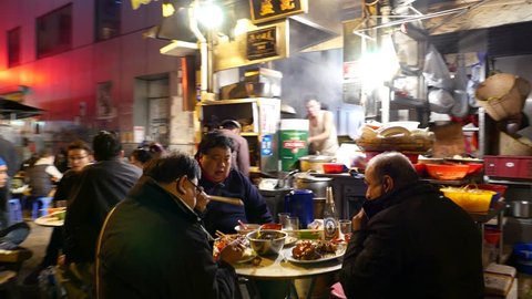 HONG KONG - FEBRUARY 10, 2015: Authentic outdoor eatery on dark urban alley, people seat and eat supper, POV walk through pedestrian street, crowded by noshery kiosks, night outside kitchen being work