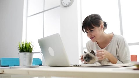 Smiling woman at home typing on a laptop and social networking, she is holding and cuddling her cat