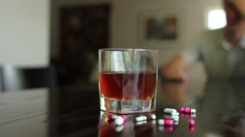 Substance abuse, pan shot of pills and alcohol out of focus man in background