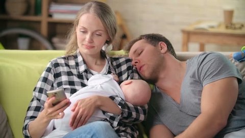 Tired young mother text messaging on a smart phone, her baby sleeping in her arms and her husband napping on her shoulder