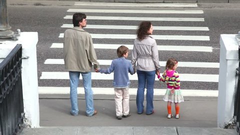 Family goes by pedestrian crossing road, parents keep childrens hands, car ride by road in front of them and behind when they passed