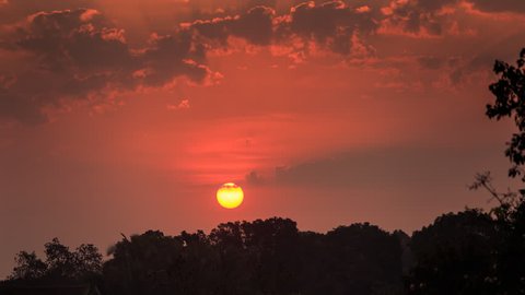 quick motion of red large sun disk among red sky and clouds above line of tree silhouettes at sunrise