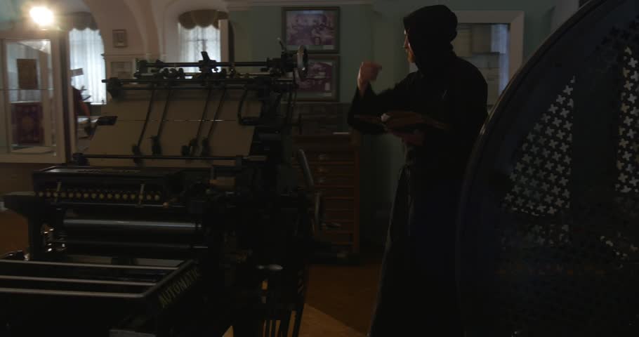 Monk, man with beard and mustache in Black Clothes, Black Hood, Holding a Book, is Walking With The Book by The Room With Brown Wooden Floor, Furniture, Windows, Pictures, Ancient Printing Tools,