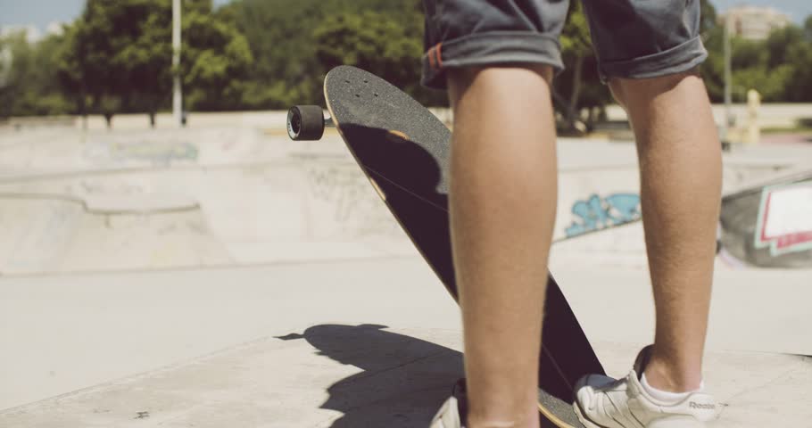 Man standing at a skate park tipping up his his longboard or skateboard with his foot as he waits on the concrete  low angle lower leg view.