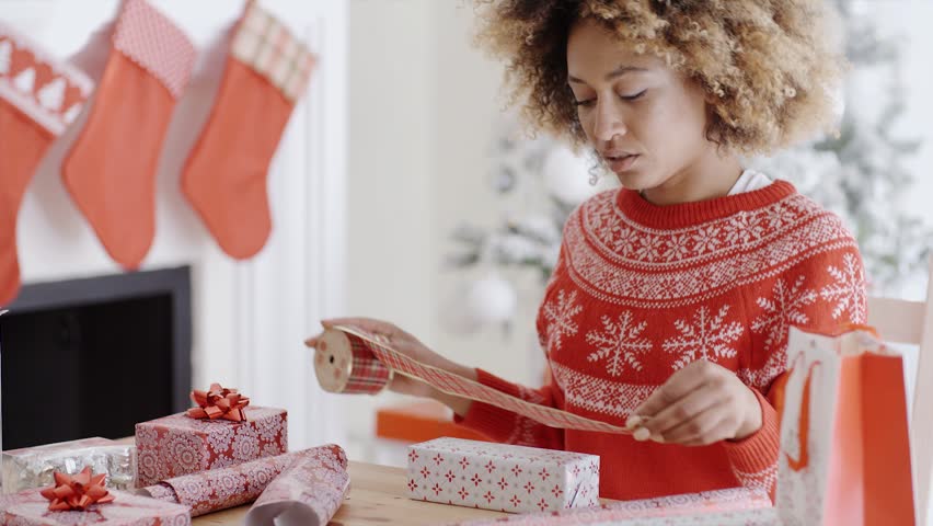 Pretty young woman sitting at a table at home wrapping Xmas presents in colorful paper in front of a decorated Christmas tree