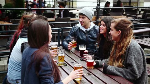 Group of friends enjoying a beer at pub in London, toasting and laughing. They are four girls and two boys in their twenties on a cloudy autumnal day - Βίντεο στοκ