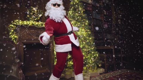 Santa Claus in sunglasses dancing and looking at the camera, tracking shot, snowflakes, christmas tree with lights and decorated fireplace in background