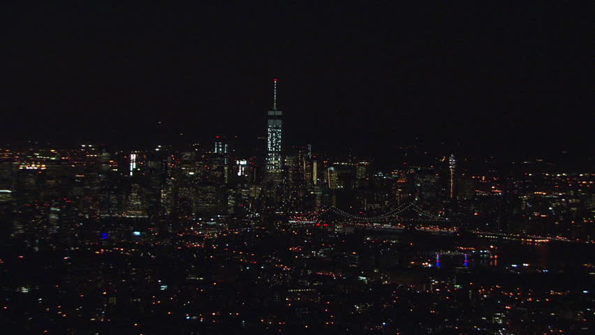 A wide aerial establishing shot at night of downtown manhattan on a clear summer night. The lights of the skyline illuminate the city sky.