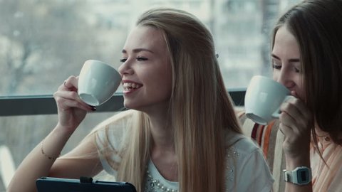 Two smiling girlfriends drink coffee and communicate