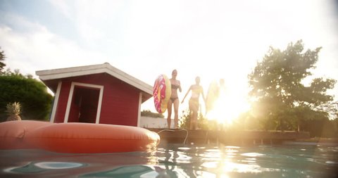 Three girls mid-air while jumping joyfully into a backyard swimming pool while holding big pool inflatables on a summer evening with sun flare in Slow Motion