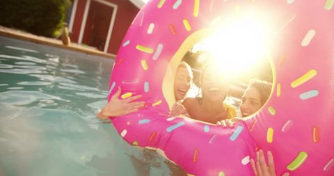 Three girls floating in a swimming pool with a pool inflatable looking like a big pink donut having fun while laughing on a summer afternoon in a backyard with sunflare