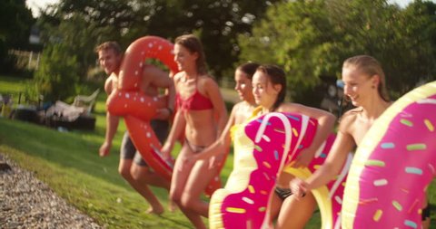 Teen friends jumping into a summer pool in Slow Motion and each of them holding a pool inflatable