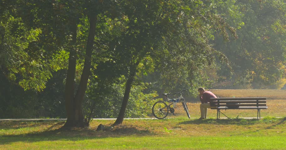 Man is Sitting on The Bench With His Arms Leaned to Knees ,Man Has a Rest, Bored Man on the Bench in Park, Backpacker ,Tourist is Sitting Alone, bicycle is standing at the bench, bench in park alley
