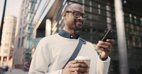 African American Man sms texting using app on smart phone in city. Handsome young businessman drinking coffee using smartphone smiling happy. Urban male professional commuting in his 20s