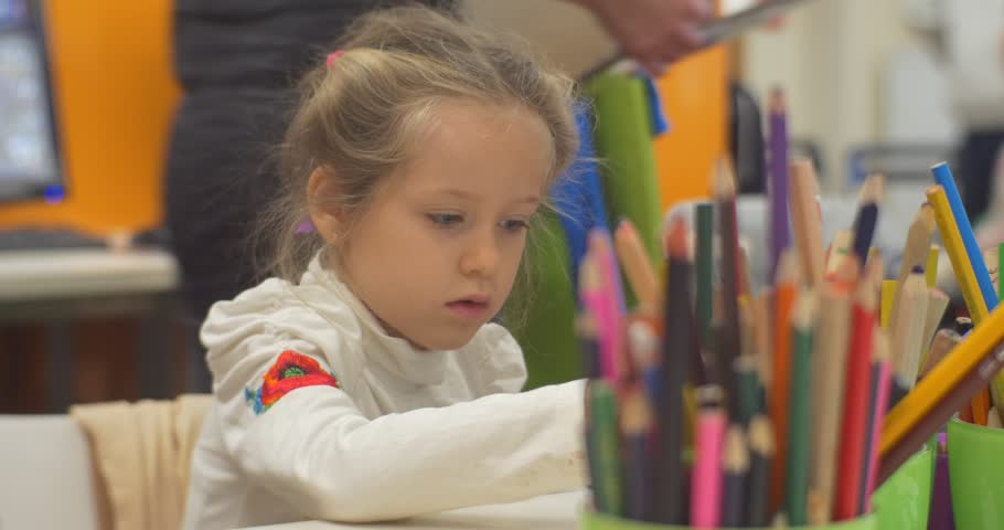 Little Blonde Girl with Concentrated Face is Sitting at The Table And Painting by Colorful Pencils, Takes a Glue, cup of pencils is on the table, Playing at Classroom of Central Library Kids are