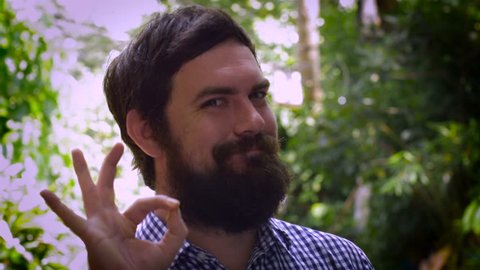 Close up portrait of a young hipster man with a full beard saying A OK with his right hand. A real life emoticon in an outdoor setting with natural lighting and a unique expression.