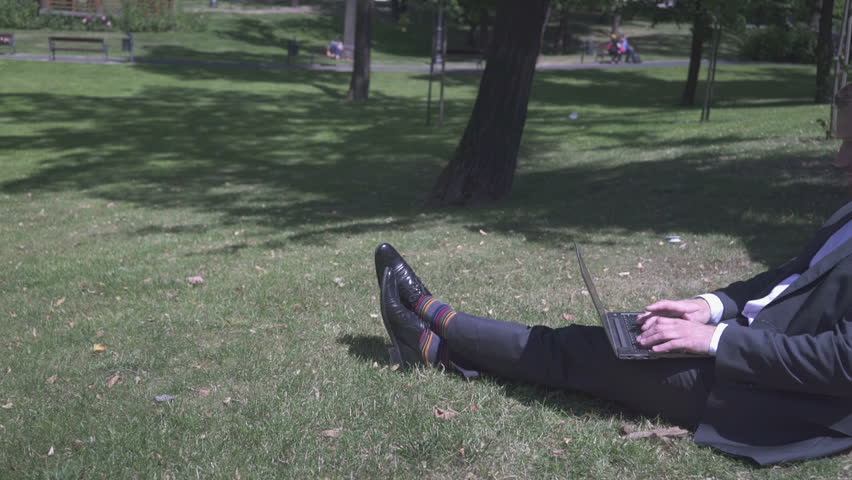 Businessman typing on laptop, sitting on ground under tree in park, pan shot.