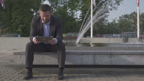 Businessman sitting on wall browsing tablet against fountain.