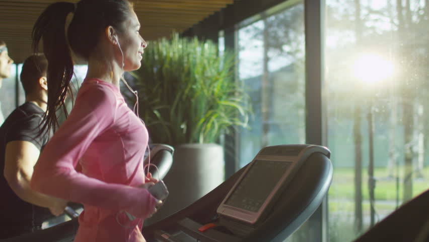 Attractive caucasian girl running on the treadmill in the sport gym with phone and earphones. Shot on RED Cinema Camera in 4K (UHD).