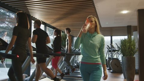 Attractive blond caucasian girl is drinking a protein shake drink while walking next to a treadmill in the sport gym. Shot on RED Cinema Camera in 4K (UHD).