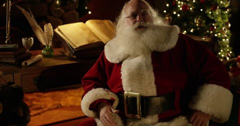 Santa Claus mouths words 'merry christmas' to camera with a perfectly decorated Christmas Home and his desk complete with Naughty/Nice list