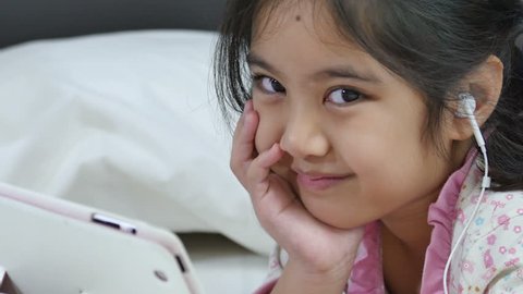 Little Asian girl in earphones using digital tablet on the bed