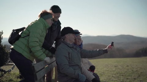 4k footage, four happy seniors taking selfies with handy on bench outside during their walk in hilly landscape
