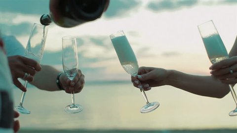 Group of friends celebrating success and pouring glasses with champagne from bottle at sunset summer sea background