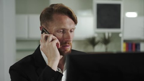 Red-haired man sitting at a computer and calling on the phone. UHD