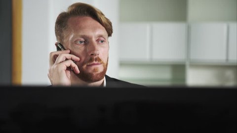 Red-haired man sitting at a computer and calling on the phone. UHD