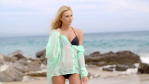 Attractive Blond Woman Wearing Black Bikini and Teal Colored Cover Up Standing with Hand in Hair on Rocky Beach