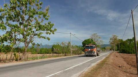 Big truck drive country road approach camera, tropical island, dry waste land, some trees, perspective view. Jalan Pantai Cenang in the Langkawi International Airport direction