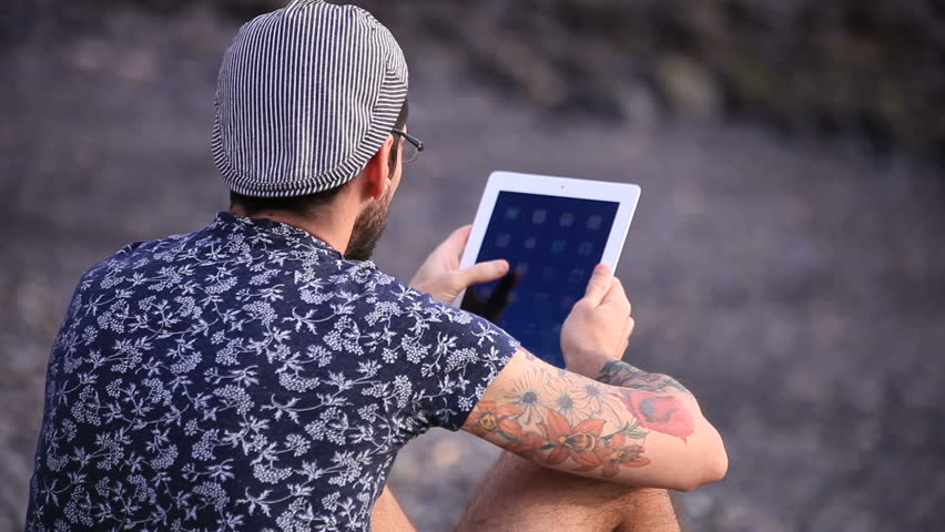A guy with flat cap and a tablet sitting on the gravel beach (close up)