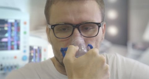 Young Man in Glasses in Manipulation Room is Breathing Through Nebulizer Mask, Hand is Holding a Mask at the Face, sick man in a ward, Parameters Displayed on the Screen, monitors of medical