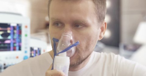Young Man in Manipulation Room is Breathing Through Nebulizer Mask, Parameters Displayed on the Screen, Steaming Nebulizer, Light Room, Hand is Holding a Mask at the Face, sick man in a ward, indoors