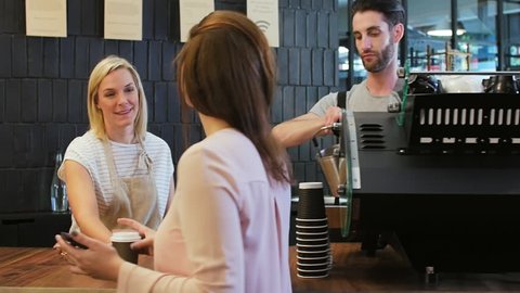 Barista at coffee machine, friendly waitress serves coffee and interacts with customer for orders in modern trendy cafe coffee shop