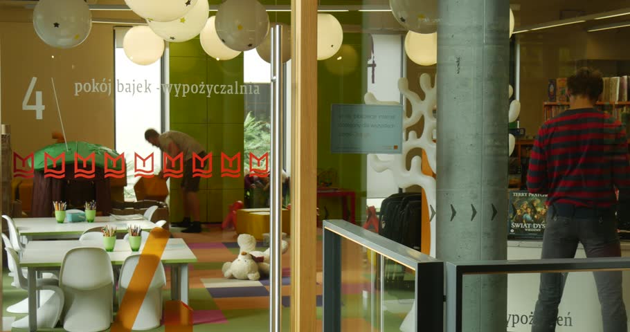 The Fairy Tale Room in John Paul II Municipal Public Library. Two men in the room. Green walls, colorful floor, high windows, white tables and chairs, lamps-balls, bookshelves with books. Sunlight