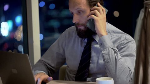 Young businessman with laptop talking on cellphone in office at night
