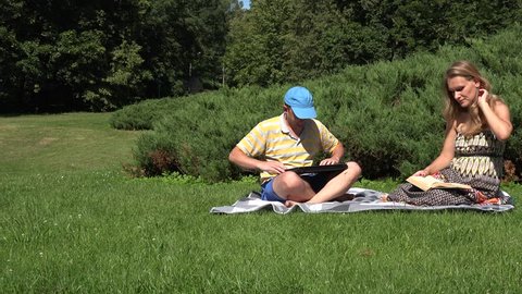 Couple man and woman lay plaid on meadow grass. Guy working with computer. Girl reading book. Family time together in park. Static shot. 4K