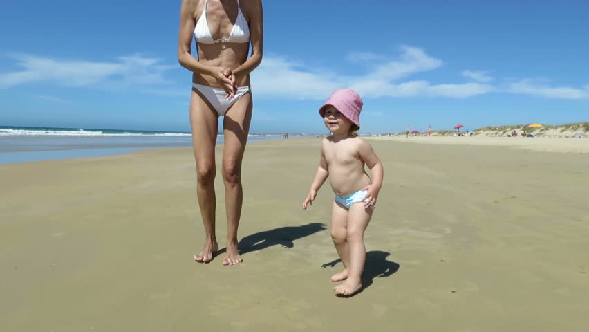 two years age baby in green swimwear with mother in white bikini dancing, jumping and playing on golden sand beach seaside with blue clear sky
